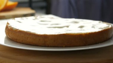 Kadın hamur üstüne krem sürüyor. Kadın eli pastanın üzerine kaşıkla beyaz krema döker ve mutfakta portakallı bisküvi veya kek hamuru sürmeye başlar.