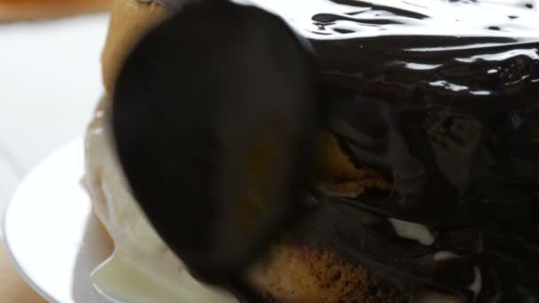 Vista de cerca. Mujer esparciendo un glaseado de hocolato en un bizcocho. La mano femenina vierte la crema con una cuchara en el pastel y comienza a extenderse en una galleta de naranja o masa de pastel de esponja en la cocina — Vídeos de Stock