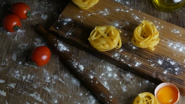 Tagliatelle ή fettuccine ζυμαρικά φωλιάζει σε ένα ξύλινο πίνακα κουζίνας δίπλα σε ένα σπασμένο κρόκο αυγού, τοματίνια, αλεύρι και ελαιόλαδο σε ρουστίκ στυλ. Εθνικό ιταλικό φαγητό — Αρχείο Βίντεο