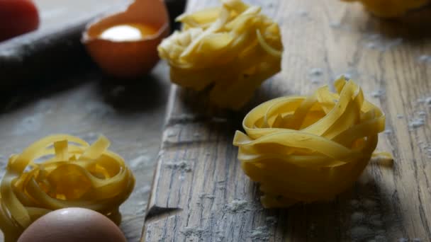 Nyers lisztből készült termékek közelről láthatók. Nemzeti olasz kaja. Tagliatelle vagy fettuccine pastas fészket egy fából készült konyhaasztalon mellett törött tojássárgája, cseresznye paradicsom rusztikus stílusban. — Stock videók