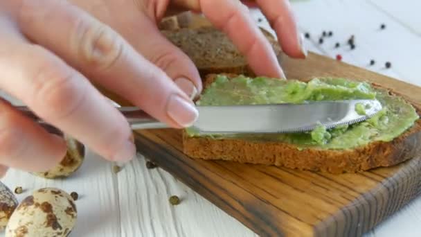 Spreid puree avocado op een geroosterd brood met een mes. Heerlijke avocado toast maken als ontbijt. Bruschetta koken met kerstomaten en kwarteleitjes. Van dichtbij gezien. Gezond veganistisch voedsel — Stockvideo