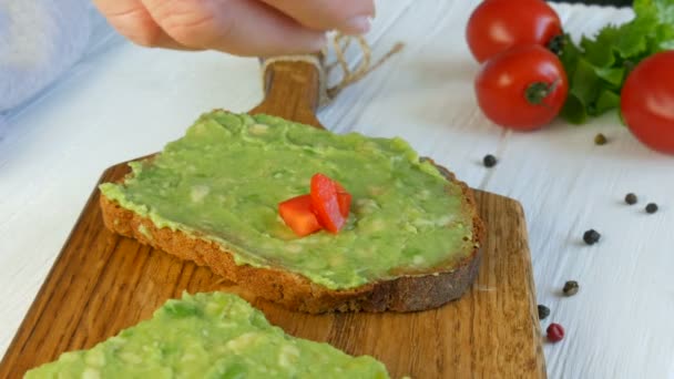 Spread mashed avocado on a toasted brown bread. Making tasty avocado toast for breakfast. Cooking bruschetta with cherry tomatoes. Healthy Vegan food — Stock Video