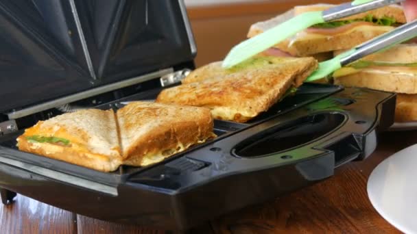 Morning breakfast in the home kitchen. Sandwiches with bacon, cheddar cheese and lettuce are fried in a special toaster or a sandwich maker. Special kitchen spatula takes fresh sandwich bread — Stock Video