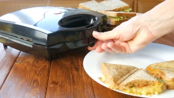 自宅のキッチンで朝の朝食。ベーコン、チェダーチーズ、レタスのサンドイッチは、特別なトースターまたはサンドイッチメーカーで揚げられています。特別なキッチンへらは新鮮なサンドイッチパンを取る — ストック動画