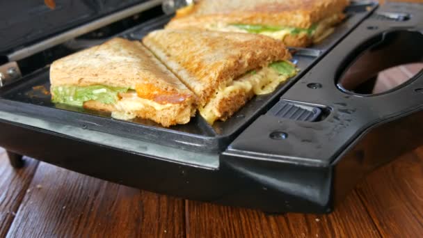 自宅のキッチンで朝の朝食。ベーコン、チェダーチーズ、レタスのサンドイッチは、特別なトースターまたはサンドイッチメーカーで揚げられています。特別なキッチンへらは新鮮なサンドイッチパンを取る — ストック動画