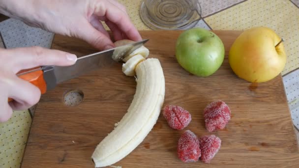 女性の手は、木製のキッチンボード上の将来のフルーツサラダやスムージーのためのナイフでバナナをカット.近くには凍ったイチゴ、黄色と緑のリンゴがあります。自宅で朝の朝食 — ストック動画
