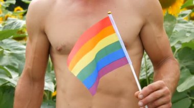 Güzel kasları olan genç bir adamın güzel vücudu gökkuşağı taşır Gey gururu Lgbt bayrağı, yaz mevsiminde açan ayçiçeklerine karşı geleneksel olmayan bir yönelimin sembolüdür.