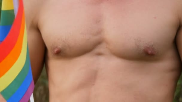 Torso é um corpo atlético atlético de um jovem bronzeado que segura em suas mãos arco-íris Orgulho gay Bandeira LGBT em suas mãos símbolo de orientação não convencional contra o fundo da natureza verde — Vídeo de Stock