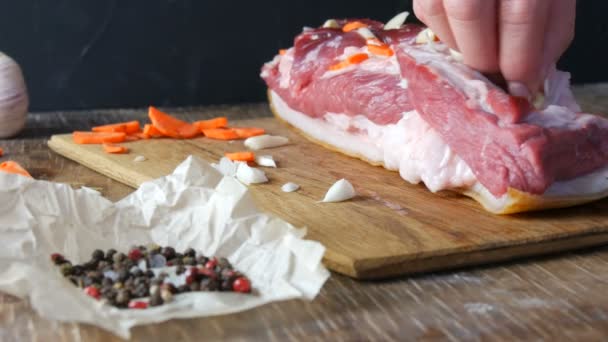 Les mains féminines poignardent avec un couteau et des assaisonnements farcis avec des tranches d'ail et de carottes un morceau de viande crue fraîche steak de porc ou de bacon dans un style rustique sur une planche de cuisine en bois. — Video