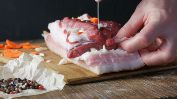 Vrouwelijke handen steken met een mes en gevulde kruiden met plakjes knoflook en wortelen een stuk vers rauw vlees varkensvlees steak of spek in een rustieke stijl op houten keukenbord. — Stockvideo