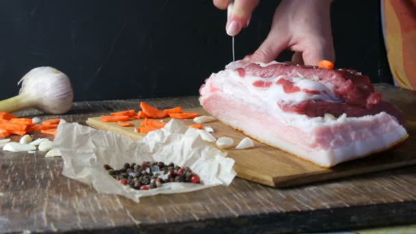 Las manos hembras apuñalan con un cuchillo y condimentos rellenos con rodajas de ajo y zanahorias un trozo de carne fresca cruda de cerdo o tocino en un estilo rústico en tablero de cocina de madera. — Vídeo de stock