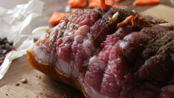Fresh juicy piece of raw meat pork steak or bacon with seasonings of pepper, garlic, carrots in a rustic style rewound with baking threads in the oven salted and spiced, garnished on wooden background — Stock Video
