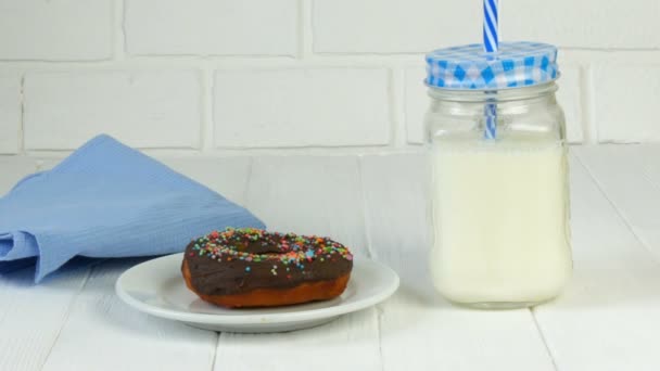 Una grande ciambella di cioccolato con polvere colorata su un piatto bianco contro un muro di mattoni accanto a una lattina di latte e un tovagliolo blu. Cibo spazzatura, diabete — Video Stock