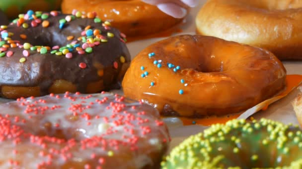 Large multi-colored fresh fried donuts in a row on a table. A beautiful donut with orange icing is sprinkled with a special colored powder for decoration of sweets — Stock Video