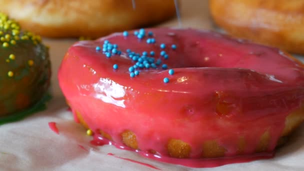 Великі різнокольорові свіжі смажені пончики поспіль на столі. Красивий пончик з рожевою глазур'ю посипається спеціальним кольоровим порошком для прикраси цукерок — стокове відео