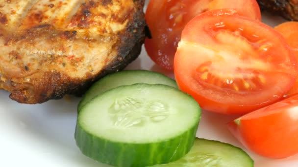 Vista de perto sobre saborosas fatias suculentas de carne de porco grelhada entrecote carne de porco ao lado de tomates cereja e legumes frescos pepinos em um prato de restaurante branco — Vídeo de Stock