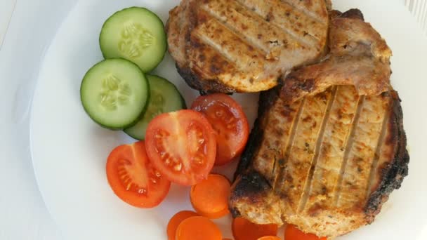 Gustose e succose fette fresche di bistecca alla griglia invitano la carne di maiale accanto a pomodorini e cetrioli di verdure fresche su un piatto bianco del ristorante. — Video Stock