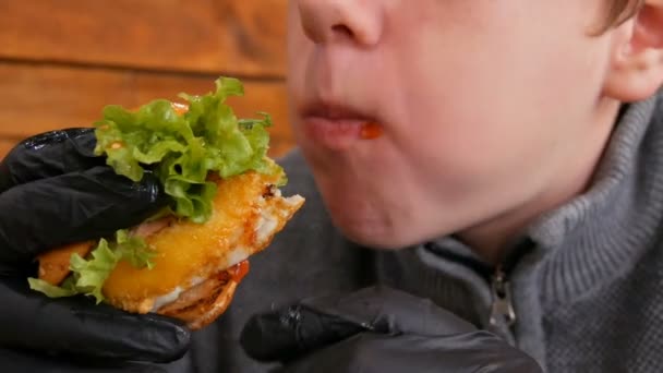 Tonårspojken i svarta gummihandskar äter en hamburgare. Läcker saftig färsk hamburgare på bordet i en snabbmatsrestaurang. salladsblad, färskt bröd, köttbit, ohälsosam sås — Stockvideo