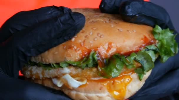 Bekijk van dichtbij een grote sappige hamburger met een ei, een tomatensnijder, salade en rode saus. Handen in speciale zwarte handschoenen houden junk food, fast food. — Stockvideo