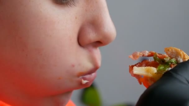 Um adolescente come grande hambúrguer suculento com um ovo, uma costeleta de tomate, salada e molho vermelho. As mãos em luvas pretas especiais mantêm a comida de lixo, comida rápida. Face e boca close up vista . — Vídeo de Stock