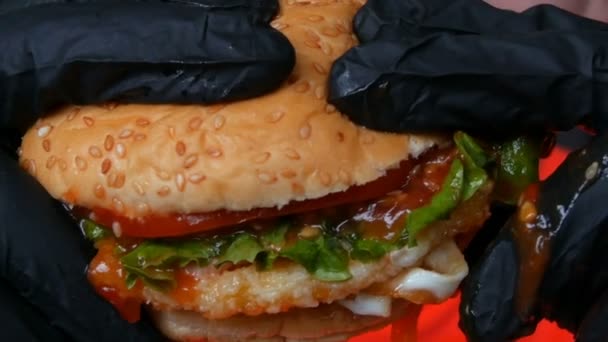 Detailní pohled na velký šťavnatý hamburger s vejcem, rajčatovou kotletkou, salátem a červenou omáčkou. Ruce ve speciálních černých rukavicích držet nezdravé potraviny, fast food. — Stock video