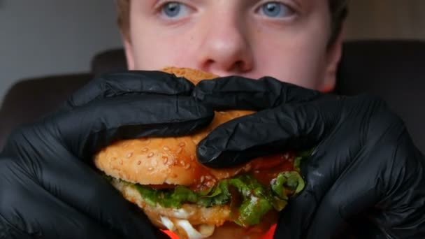 Подросток ест большой сочный гамбургер с яйцом, томатной котлеткой, салатом и красным соусом. Руки в специальных черных перчатках держат вредную пищу, фаст-фуд. Face and mouth close-up view . — стоковое видео