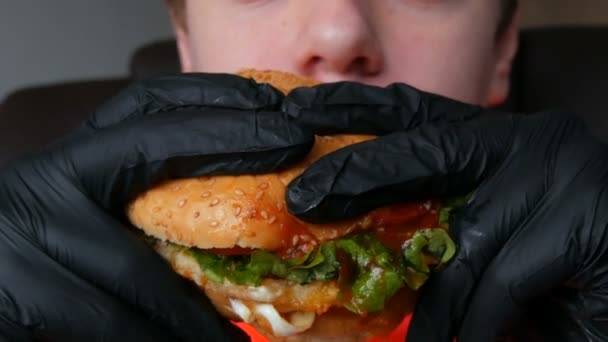 En tonåring äter en stor saftig hamburgare med ägg, tomatpuré, sallad och röd sås. Händerna i speciella svarta handskar håller skräpmat, snabbmat. Ansikte och mun närbild. — Stockvideo