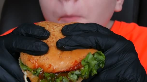 Um adolescente come grande hambúrguer suculento com um ovo, uma costeleta de tomate, salada e molho vermelho. As mãos em luvas pretas especiais mantêm a comida de lixo, comida rápida. Face e boca close up vista . — Vídeo de Stock