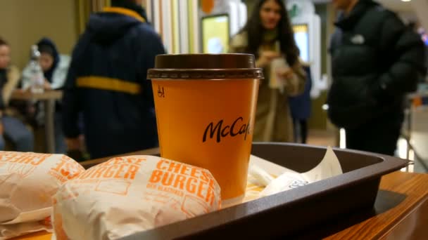 Bécs, Ausztria - 2019. december 19.: Egy pohár tejeskávé és egy pár sajtburger tálcán McDonaldwe-ban, a pénztárgép nélküli megrendelések digitális képernyőinek hátterében — Stock videók