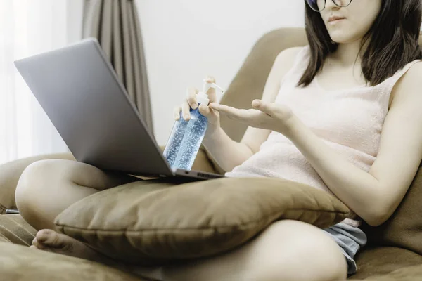 Young woman lady sit at sofa and using laptop computer put pillow and using algohol gel or hand sanitizer for wash her hand. Concept work from home and social distancing from covid-19
