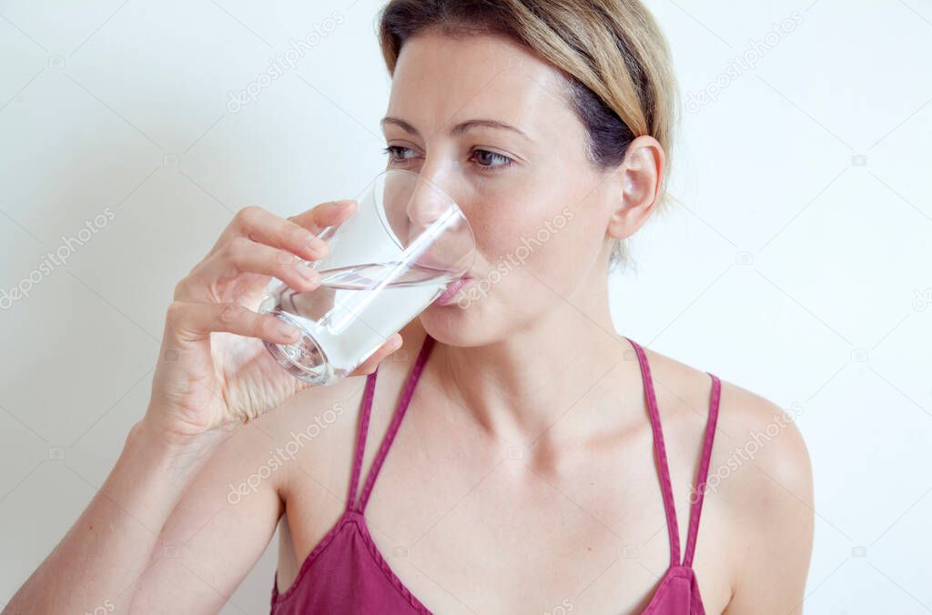Beautiful girl drinks clear water from a glass cup on a light background. Healthy lifestyle concept.