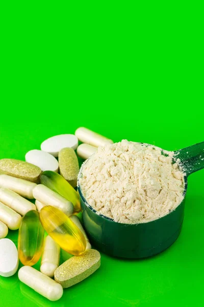Vegyes természetes étrend-kiegészítő tabletták és fehérje por műanyag kanállal zöld háttér. — Stock Fotó