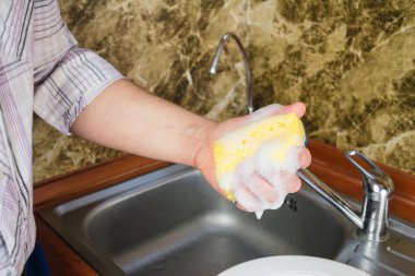 Mutfakta bulaşıkları köpükle yıkamak için sünger tutan adam.
