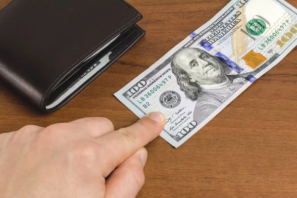 Adam, ahşap arka planda, yakın planda, para ve finans konseptinde 100 dolarlık banknotun üzerine parmağını koydu.