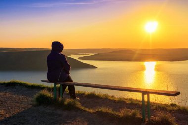 Bankta oturan ve Dinyester nehrinde muhteşem günbatımını izleyen bir kadın. Güneş suya yansıyor.