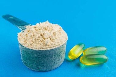Plastik kaşıkta protein tozu ve mavi arka planda omega 3 kapsülleri, spor beslenmesi.