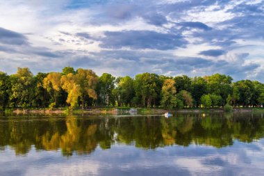Ağaçlar ve bulutlar nehirde yansıyor, insanlar suda küçük bir katamaran üzerinde dinleniyor..
