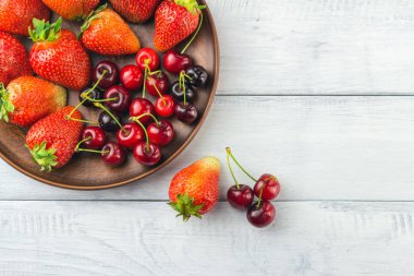 Taze organik böğürtlen, çilek ve kiraz hasadı beyaz ahşap zemin üzerinde kahverengi tabakta, üst manzara.