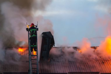 İtfaiyeci ya da itfaiyeci, yangın ateşini evin çatısından dumanla söndürüyor..
