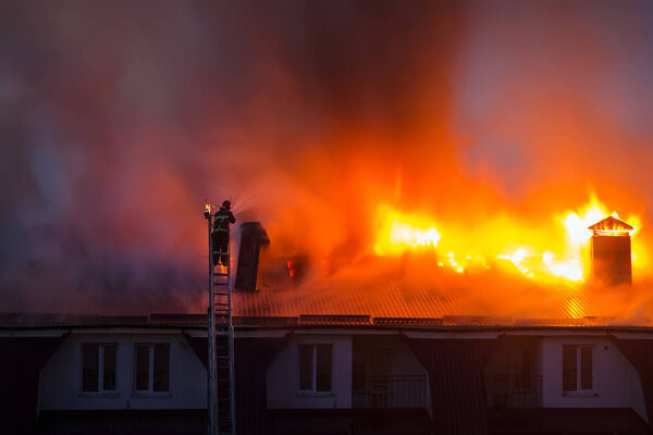 Burning fire flame with smoke on the apartment house roof in the city, firefighter or fireman on the ladder extinguishes fire.