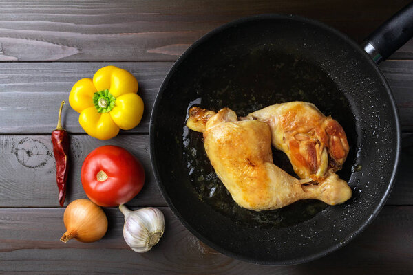 Two roasted chicken legs in black frying pan with vegetables and spices on dark wooden planks, top view.