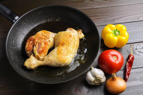 Two roasted chicken legs in black frying pan with vegetables and spices on dark wooden planks.
