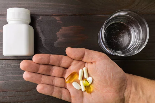 Hand hold vitamin complex and dietary supplement, capsules of omega 3, glucosamine, calcium pills, white bottle and glass of water at wooden table, top view.