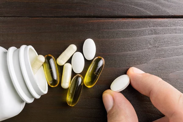 Hand takes white calcium pills, complex of vitamins and dietary supplement, yellow capsules omega 3, white capsule of glucosamine and plastic bottle at wooden table, top view.