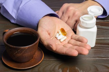 Erkek elinde vitamin kompleksi ve beslenme takviyesi, omega 3 kapsülleri, glukozamin, kalsiyum hapları, koyu ahşap masada bir fincan sade kahve var..