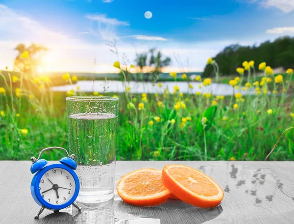 Glass of fresh cold water with oranges on the table among of picturesque the green flower meadows. Homemade juice with fresh citruses on the background of nature. The freshness of a summer morning.