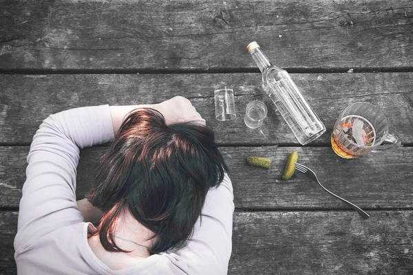 Chica joven borracha durmiendo en la vieja mesa de madera después de la fiesta. Botella de vodka y pepinillos, chupitos y jarra de cerveza. Alcoholismo femenino. Resaca. Dependencia al alcohol. Mal hábito . — Foto de Stock