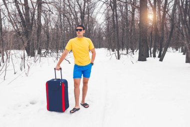 Kış yaz için seyahat. Güneş gözlüğü, parmak arası terlik, t-shirt ve şort turist bavulla genç sevimli adam karda orman geçer. Rüya tatil, deniz, sıcak egzotik ülke.