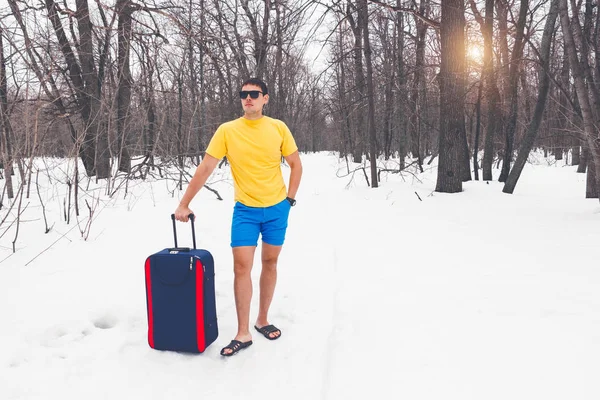 Kış yaz için seyahat. Güneş gözlüğü, parmak arası terlik, t-shirt ve şort turist bavulla genç sevimli adam karda orman geçer. Rüya tatil, deniz, sıcak egzotik ülke.