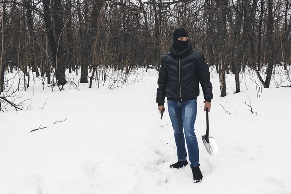 Murderer in the black mask with gun and shovel leaves place of the crime, going through the snow in the dark forest at the evening.